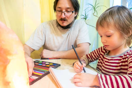 Bearded man in grey t-shirt and cute three - year - old boy drawing pictures with colored wax...