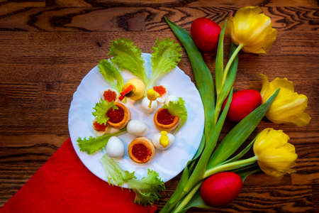 red eggs are lying on the table among yellow tulips tied with a scarlet ribbon. A plate with quail eggs, caviar nests and a chicken figurine made of yolks is the concept of an Easter dishの写真素材