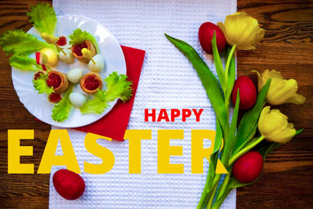 red eggs are lying on the table among yellow tulips tied with a scarlet ribbon. A plate with quail eggs, caviar nests and a chicken figurine made of yolks is the concept of an Easter dishの写真素材