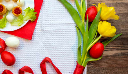 red eggs are lying on the table among yellow tulips tied with a scarlet ribbon. A plate with quail...