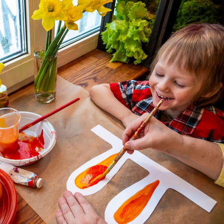 womens speckled hands teach boy draws paper rabbit ears. Preparation for Easter: a child paints paper rabbit ears with paints in the kitchen among fresh greenery, flowers and painted eggsの写真素材
