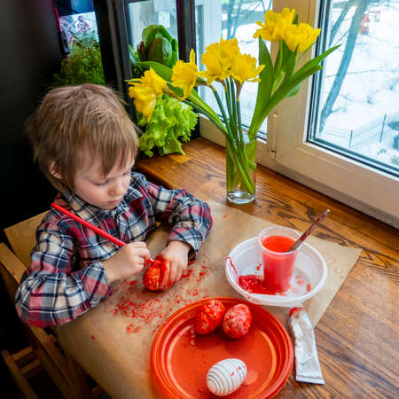 funny kid in a red plaid blouse paints artificial Easter eggs red on a wooden table among fresh greenery. preparation for the bright Easter holidayの写真素材