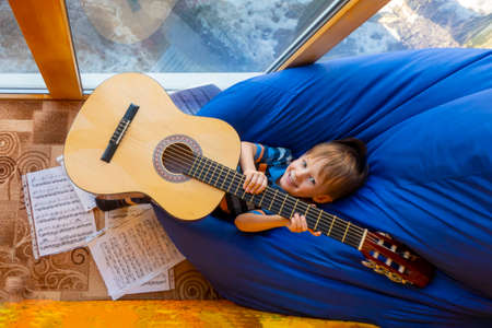 small child is playing with a guitar, sitting on a blue bag chair on a glazed sunny balcony in a...