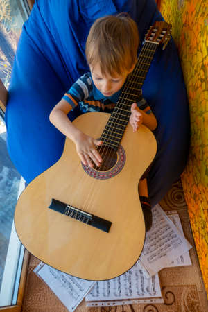 small child is playing with a guitar, sitting on a blue bag chair on a glazed sunny balcony in a snow-covered cityの写真素材
