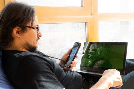 serious man sits in a blue bean bag chair and uses a laptop indoors. home office on the balcony. freelancing and remote work conceptの写真素材