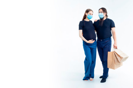 young pregnant woman and her husband with shopping bags in medical masks. safety from colds flu and coronavirus on white background. Pregnancy shopping concept happy young family with shopping bagsの写真素材