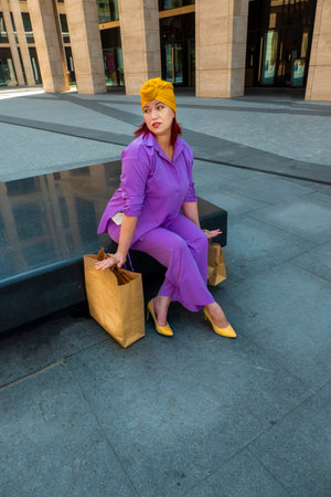 woman with craft shopping bags in the city. The concept of fashionable Consumerism and ecology. Adult woman in a purple suit with red and yellow accents enjoys shopping. banner with copy spaceの写真素材