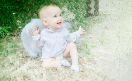 Fairy tale concept. Little toddler girl wearing beautiful dress with angel wings in the forest or park, spring day, white blooming trees, poplar fluff cloudsの写真素材