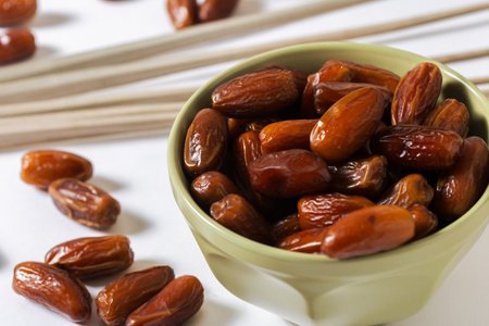 Fresh organic dates in a green bowl with scattered dates and palm leaves on a white background,...