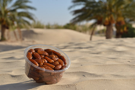 A close-up of fresh, glossy dates in a transparent plastic container placed on golden desert sand,...