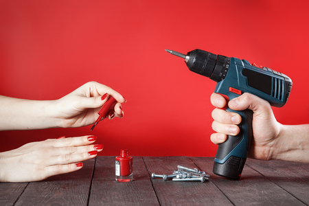 Concept of confronting domestic male and female worries. Female hand paints fingernails, and man's hand holds electric screwdriver. Close-up on wooden table. Red background.の写真素材