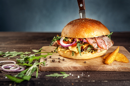 Tasty pork hamburger on dark wooden board pierced with knife, surrounded by french fries, raw meat and black pepper. Free space for textの写真素材