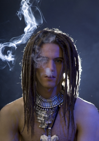 a young stylish man with dreadlocks and a lot of silver jewelry smoking a cigarette, blue filter effect の写真素材