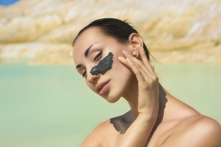 Woman with black Clay Facial Mask. Beauty and Wellness. Spa Outdoor, Beautiful Young Woman with natural Dead Sea Facial Mask on her Face near the Lake with Blue and White Cosmetic Healing Clayの写真素材