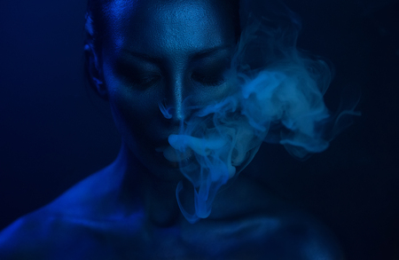 Halloween Vape Party, Nightlife. Beautiful Sexy Young Woman with glamorous mystical makeup vaping in Nightclub, exhaling smoke. Girl smoking vaporizer in Club. Blue mystic smokeの写真素材