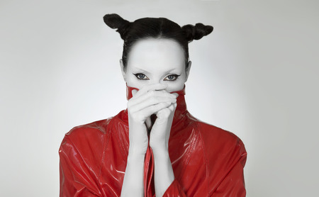 Creative glamor. Young Woman In fashionable red Long lacquer raincoat. Crazy Fashion Make-up, Accessory and Creative Hairstyle. Stylish demon, Total black look. Girl with white faceの写真素材
