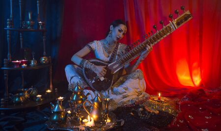 Beautiful young indian woman in traditional Sari clothing with Oriental Jewelry Playing the Sitar. Indian woman Playing night Raga the Sitar, Eastern Music. Musical Candlelight Party in India.の写真素材