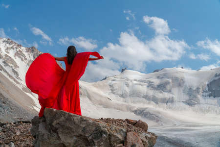 The girl stands on a stone pedestal in a developing red dress against the background of clouds, a...