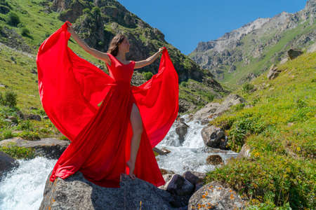 A girl in a red flowing dress stands near a mountain river. A gentle combination of blue sky, green...