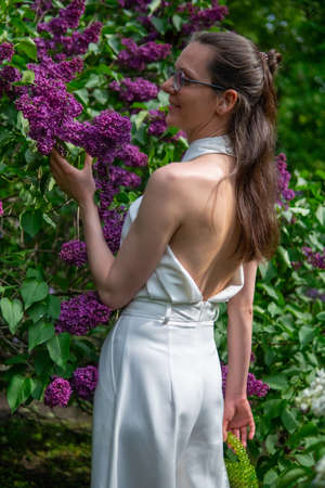 A young girl smiling next to lilac bushes in a white jumpsuit with an open backの写真素材