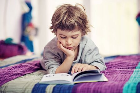 Cute little boy lying on bed reading bookの写真素材