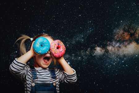 Happy cute girl is having fun played with donuts on cosmos background wall. Bright photo of a child. Colored donutsの写真素材