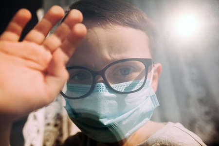 a child in quarantine because of a virus sits at home in a mask and looks out the window on a sunny day.Coronavirus and Air pollution pm2.5 concept. COVID-19の写真素材