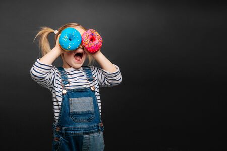 Happy cute girl is having fun played with donuts on black background wall. Bright photo of a child. Colored donutsの写真素材