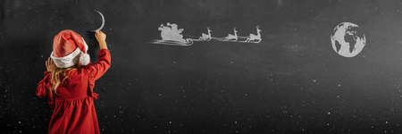 Girl in santa claus hat draws christmas drawing on the blackboardの写真素材