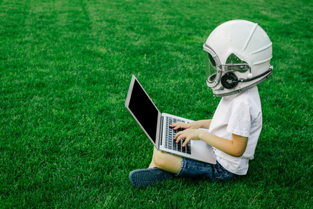 A child sits on the grass in an astronaut's helmet and studies on a laptop online enjoying nature.の写真素材