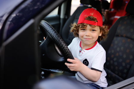 child with curly hair and a red cap sits behind the wheel of a car. baby boy grimaces in car windowの写真素材