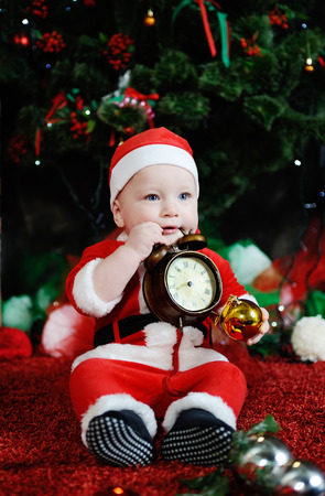 Child dressed as Santa Claus gnawing teeth clock alarm clock. Child dressed as Santa sitting on the background of the Christmas treeの写真素材