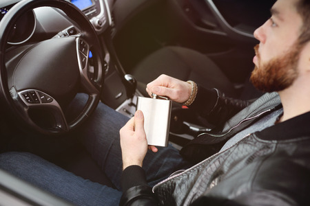 Young man driving a car with an iron flask with alcohol. Danger on the road. Bad habit. Drunk Driver.の写真素材