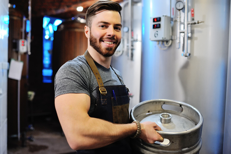 Male brewer with a metal beer barrel on the background of the brewery