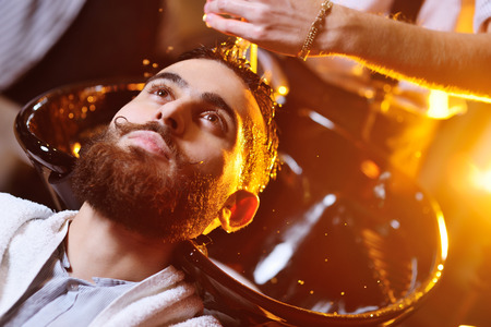 Barber or hairdresser washes the head of the client - a young handsome man with a beautiful beard and mustache.の写真素材
