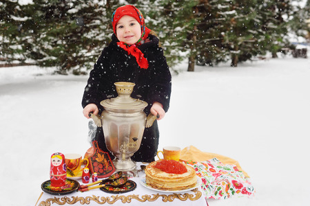 child girl in a fur coat and in a scarf in Russian style holding a large samovar in the hands of pancakes with red caviar, matryoshkas and utensils in Khokhloma style. Maslenitsa, Russia, winterの写真素材