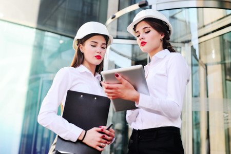 Two young pretty business women industrial engineers in construction helmets with a tablet in hands on a glass building background. Construction plan, architect, designer, successfulの写真素材