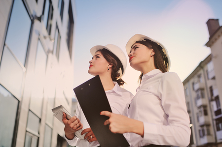 Two young pretty business women industrial engineers in construction helmets with a tablet in hands on a glass building background. Construction plan, architect, designer, successfulの写真素材