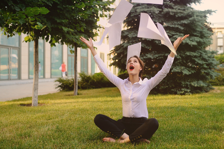 Girl in business office clothes sitting on grass throwing office documents or white sheets of paperの写真素材