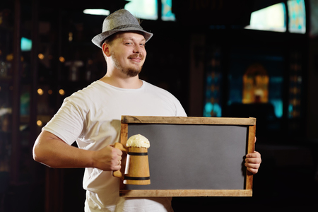 cute cheerful fat man in a Bavarian hat holding a chalkboard or a plate and a glass of beer in front of a pub during the celebration of the Oktoberfest.の写真素材
