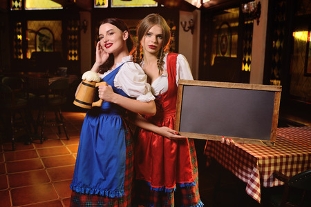two young attractive girls in traditional Bavarian clothes with chalk Board and a glass of beer in their hands on the background of the pub during the Oktoberfest celebration.の写真素材