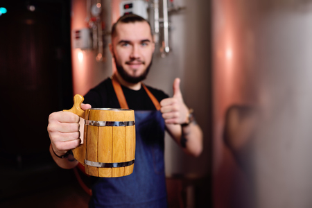 young bearded attractive male brewer with wooden beer mug in hand on background of breweryの写真素材