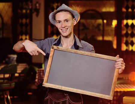 a young attractive male waiter in Bavarian clothes and wearing a hat with a feather holding a chalkboard or a plaque in his hands. Copy spaceの写真素材