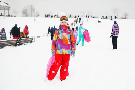 little girl in colored outerwear with a sledge child carriers in hands on the background of snow slope. Winter entertainmentの写真素材