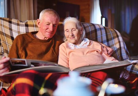 an elderly married couple-a man and a woman in a cozy house sitting on the sofa and wrapped in a warm plaid plaid hug each other and smile while viewing the family photo album.の写真素材