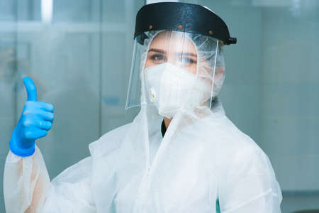 young attractive female PCR lab researcher shows a thumbs up sign class in protective suit, helmet, respirator, blue rubber gloves.の写真素材