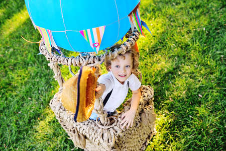 a little boy with curly hair in a straw hat sitting in a basket of a blue balloon smiles against the...