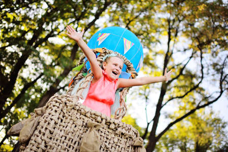 a little girl in pink clothes smiles sitting in a basket of a blue balloon against a background of sunlight and greenery.の写真素材