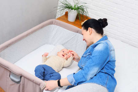 mother plays with a baby who is lying in a side-bed for a newborn or in a modern playpenの写真素材