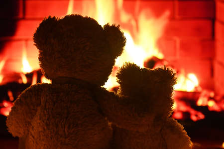 two teddy bears are sitting hugging and looking at the fireplace flame.の写真素材